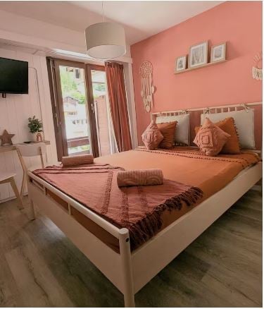 Cozy bedroom with a double bed, nightstand, chair, and decorative wall hanging. Large window with a view and a small table with a plant.