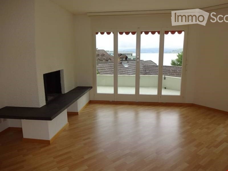 Empty room with wooden flooring, large glass doors leading to a balcony, and a built-in fireplace with a bench.