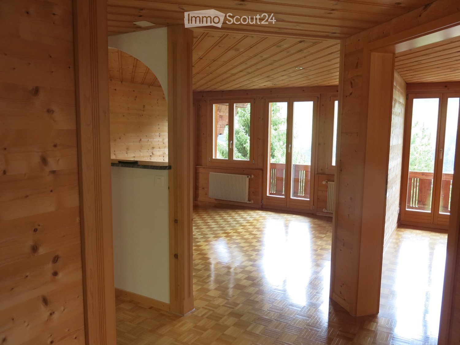 Bright, open wooden-framed room with checkered wooden flooring, large windows, and sliding glass doors leading to a balcony.