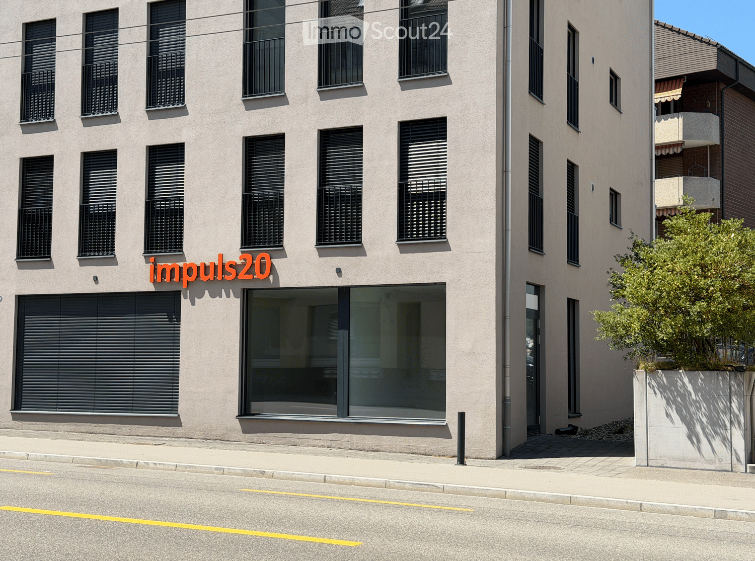 Modern multi-story building with large windows, black shutters, and a glass entrance. The facade is light-colored with a clean, contemporary design.