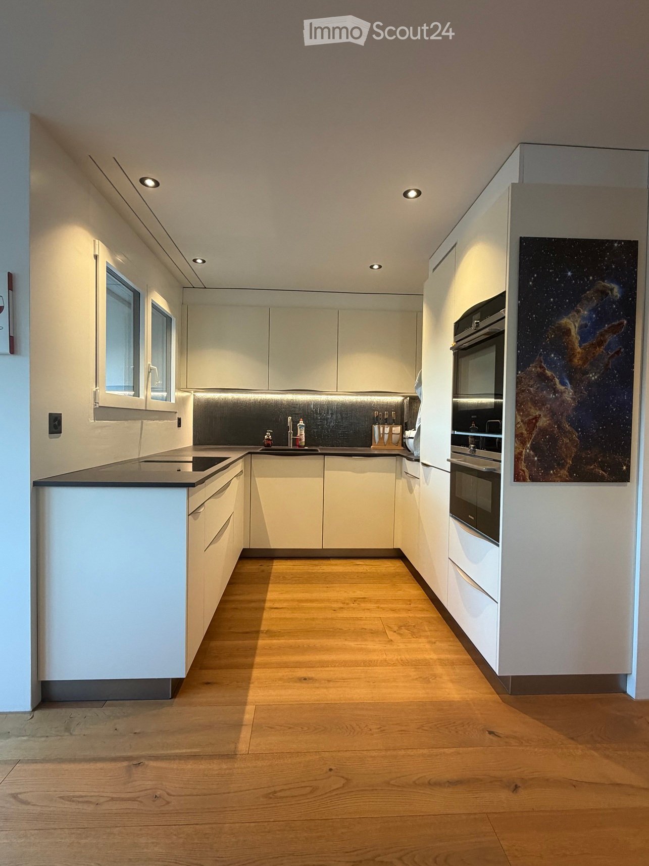 Modern kitchen with white cabinets, wooden flooring, and a sleek black countertop. Features include an integrated oven, a sink with a modern faucet, and a large painting on the wall.
