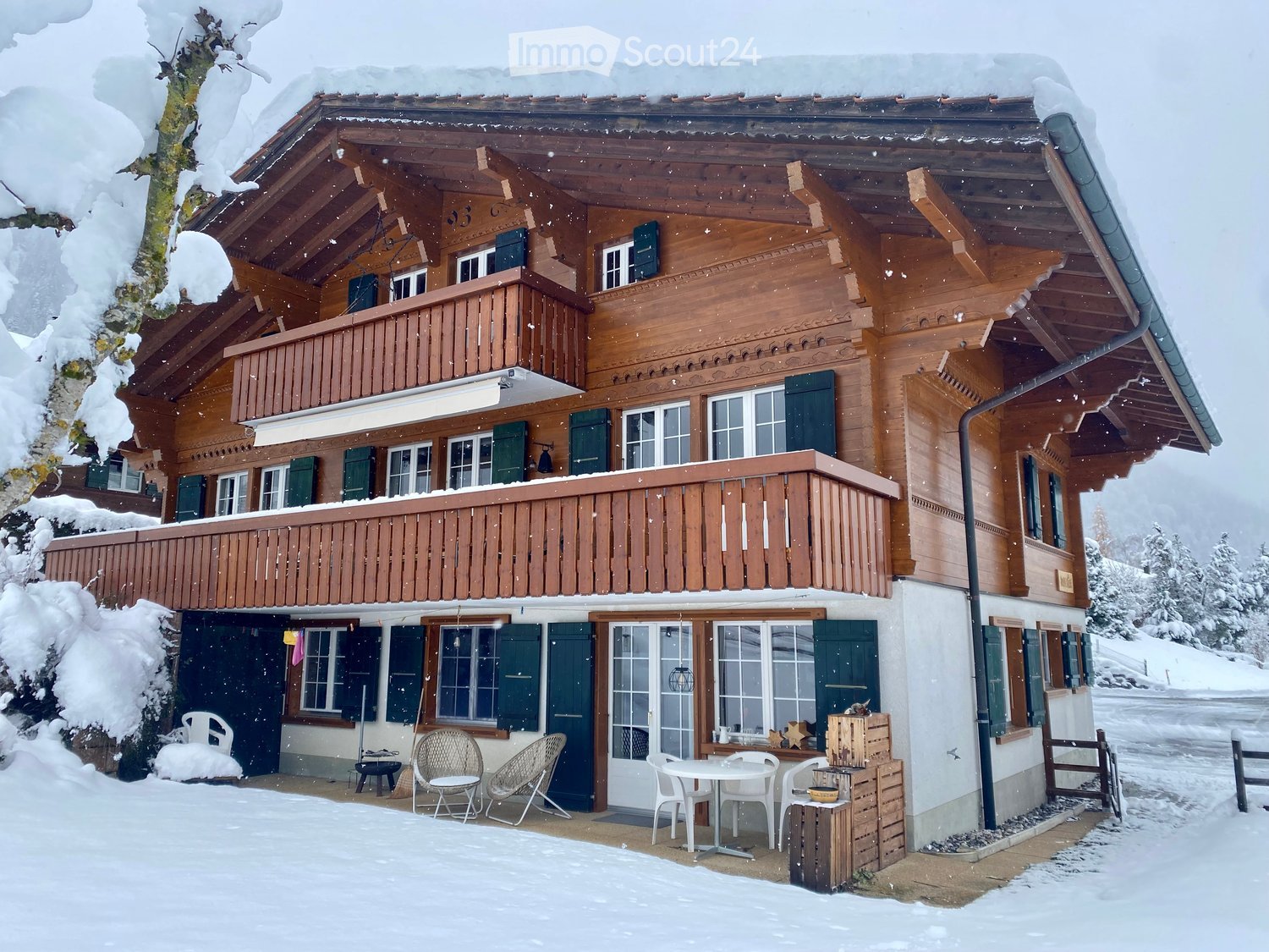 A large wooden house with multiple levels, green shutters, and a balcony, surrounded by snow-covered trees and a snowy landscape.