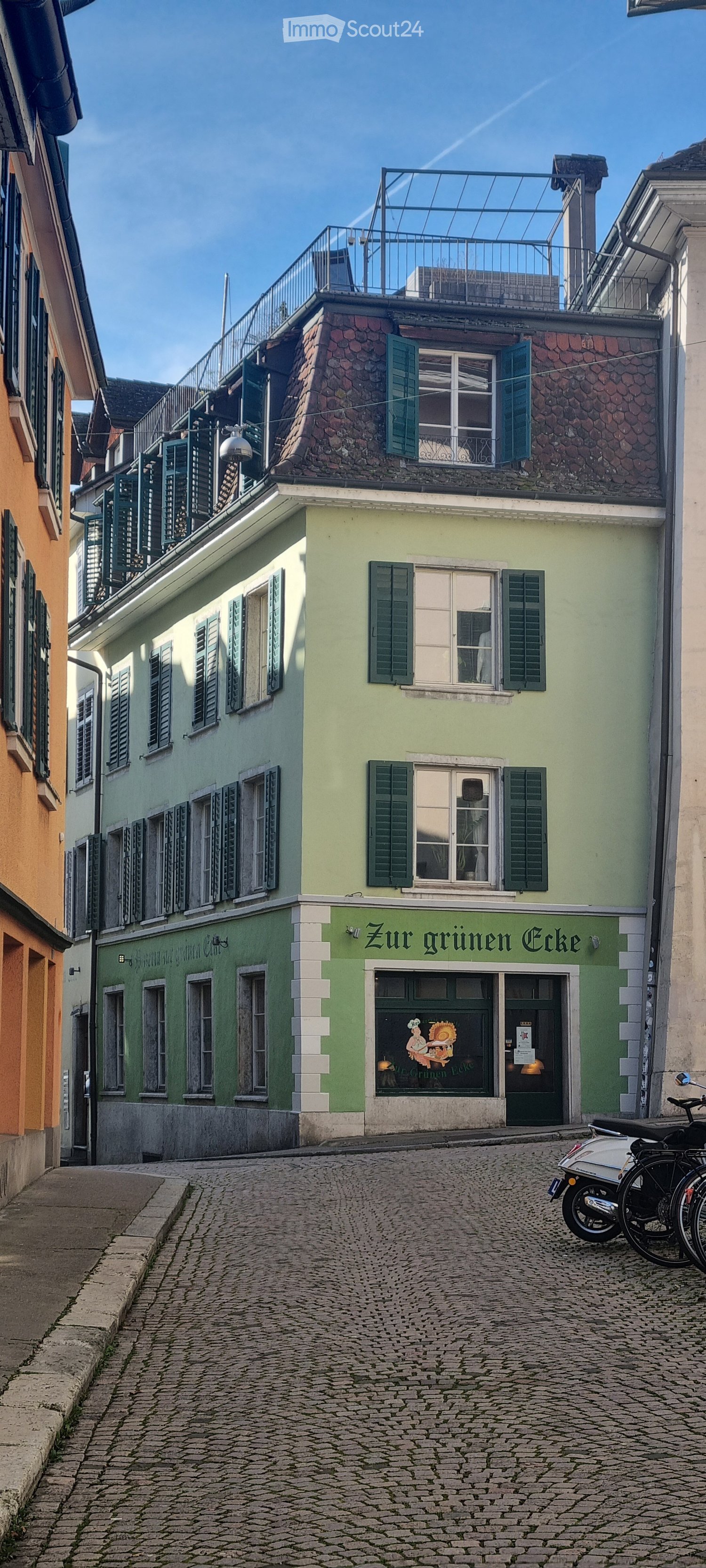 Multi-story building, green and beige exterior, many windows, metal railing on top floor, tiled street