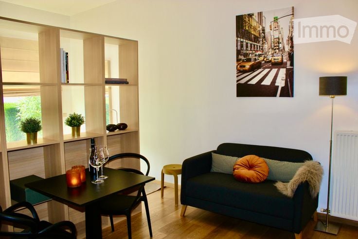 A modern, well-lit living room with a dark blue sofa, a small black dining table with chairs, a floor lamp, and built-in wooden shelves holding potted plants and decorative items.