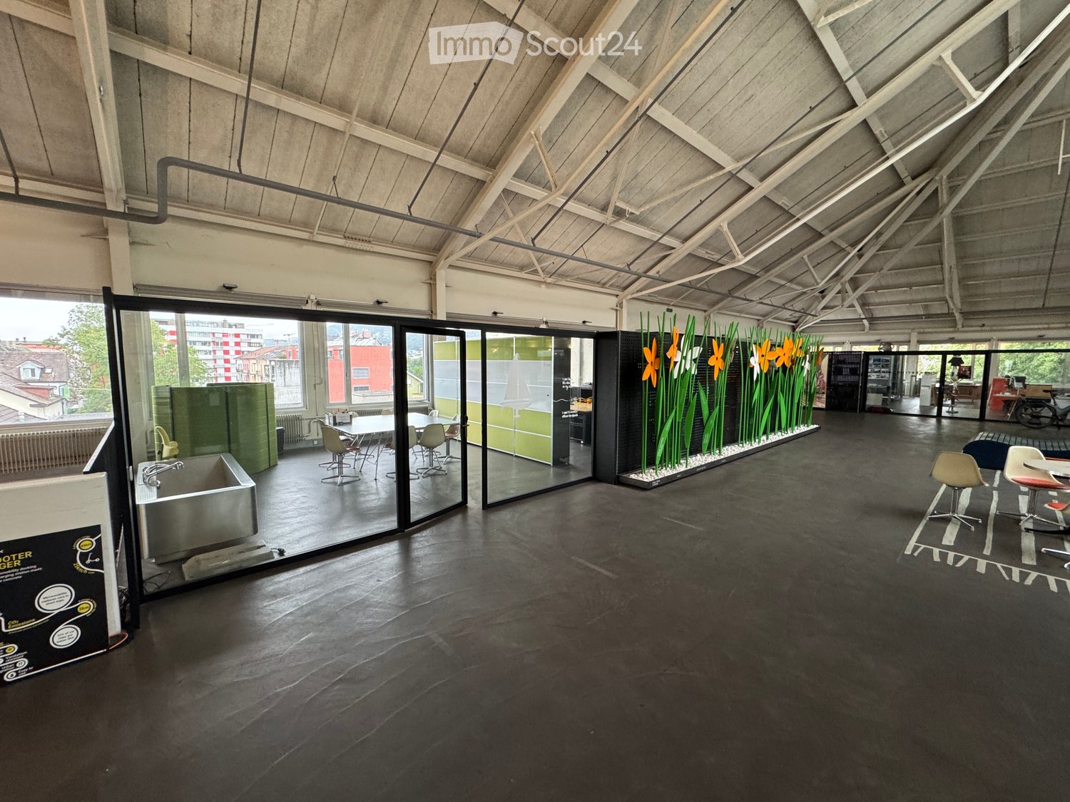 Modern, open-plan space with dark concrete floors, large glass partitions, and contemporary furniture including tables, chairs, and stools. Features a sink and decorative floral arrangement.