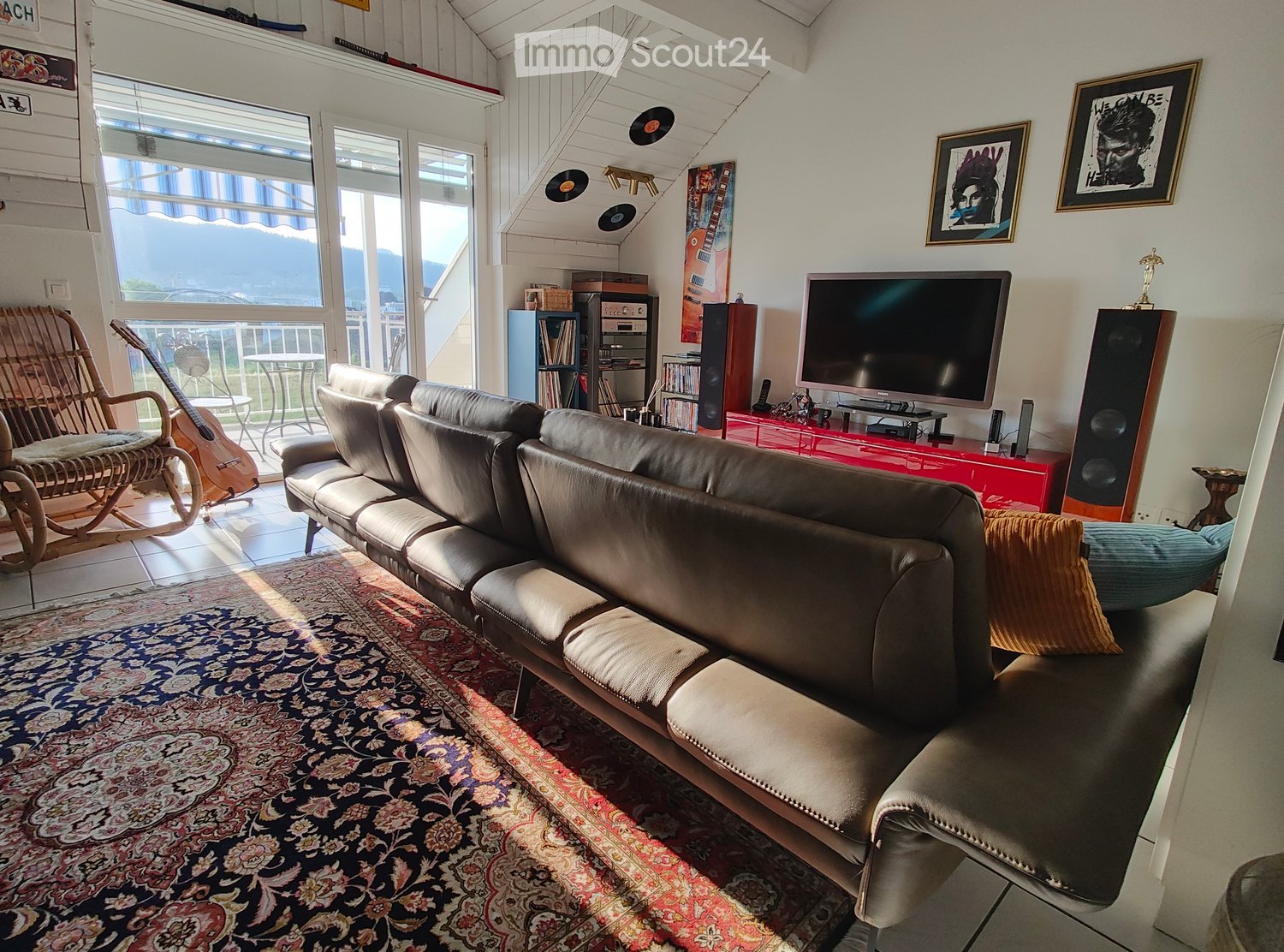 Modern living room with large windows offering a scenic view, featuring a leather sofa, ottoman, wicker chair, guitar, bookshelf, television, speakers, and framed artwork.