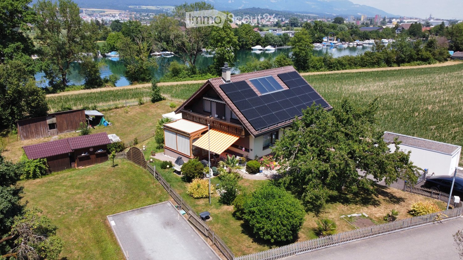 A two-story house with solar panels on the roof, surrounded by a well-maintained garden, a small wooden shed, and a view of a lake and distant mountains.