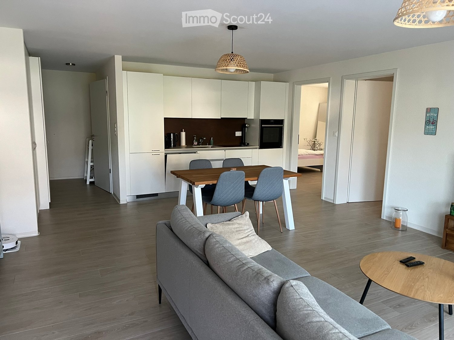 Modern open-plan living and dining area with a grey sofa, coffee table, dining table with chairs, and a fully equipped kitchen with white cabinets and stainless steel appliances.