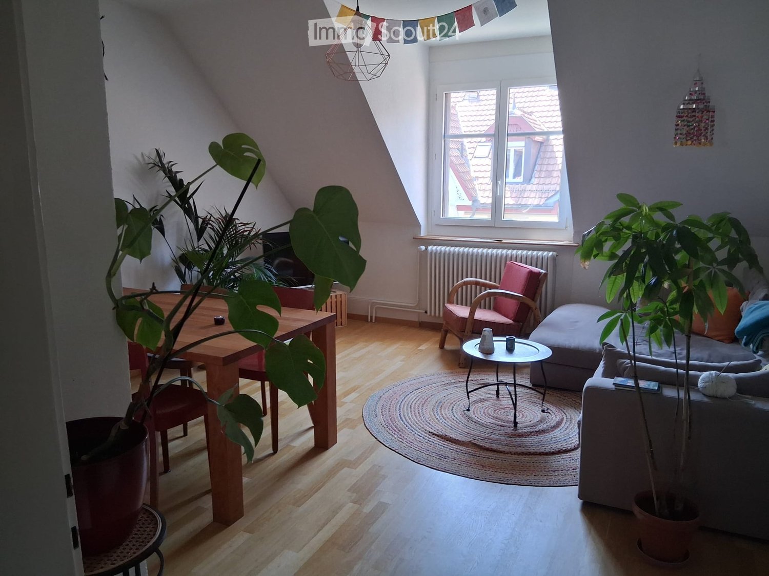 Bright living room with wooden flooring, large window, sofa, armchair, coffee table, dining table, and potted plants.