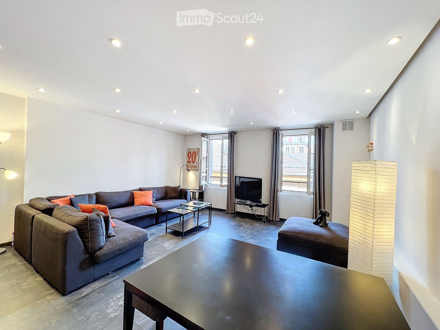 Modern living room with grey sectional sofa, coffee table, ottoman, and a television on a stand. The room features large windows with curtains and recessed ceiling lighting.