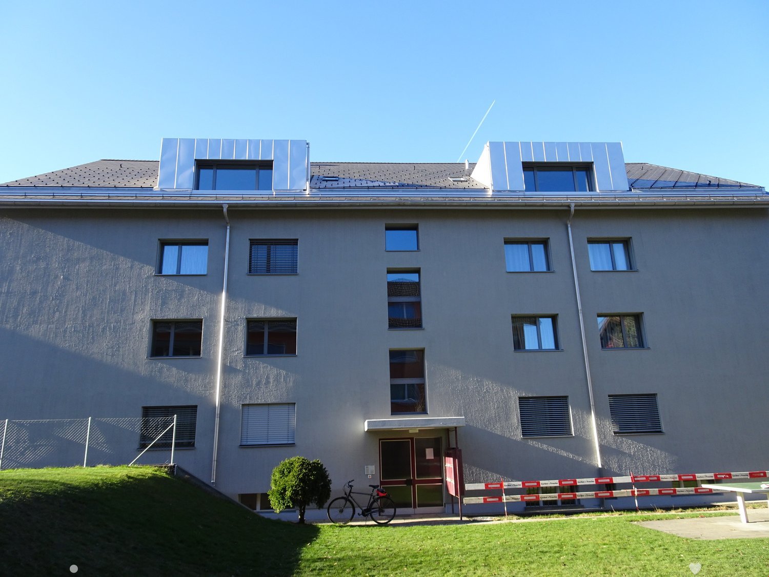 Modern two-story building with light gray facade, multiple windows, and a raised entrance area. Features a small garden with a bicycle and a table in front.