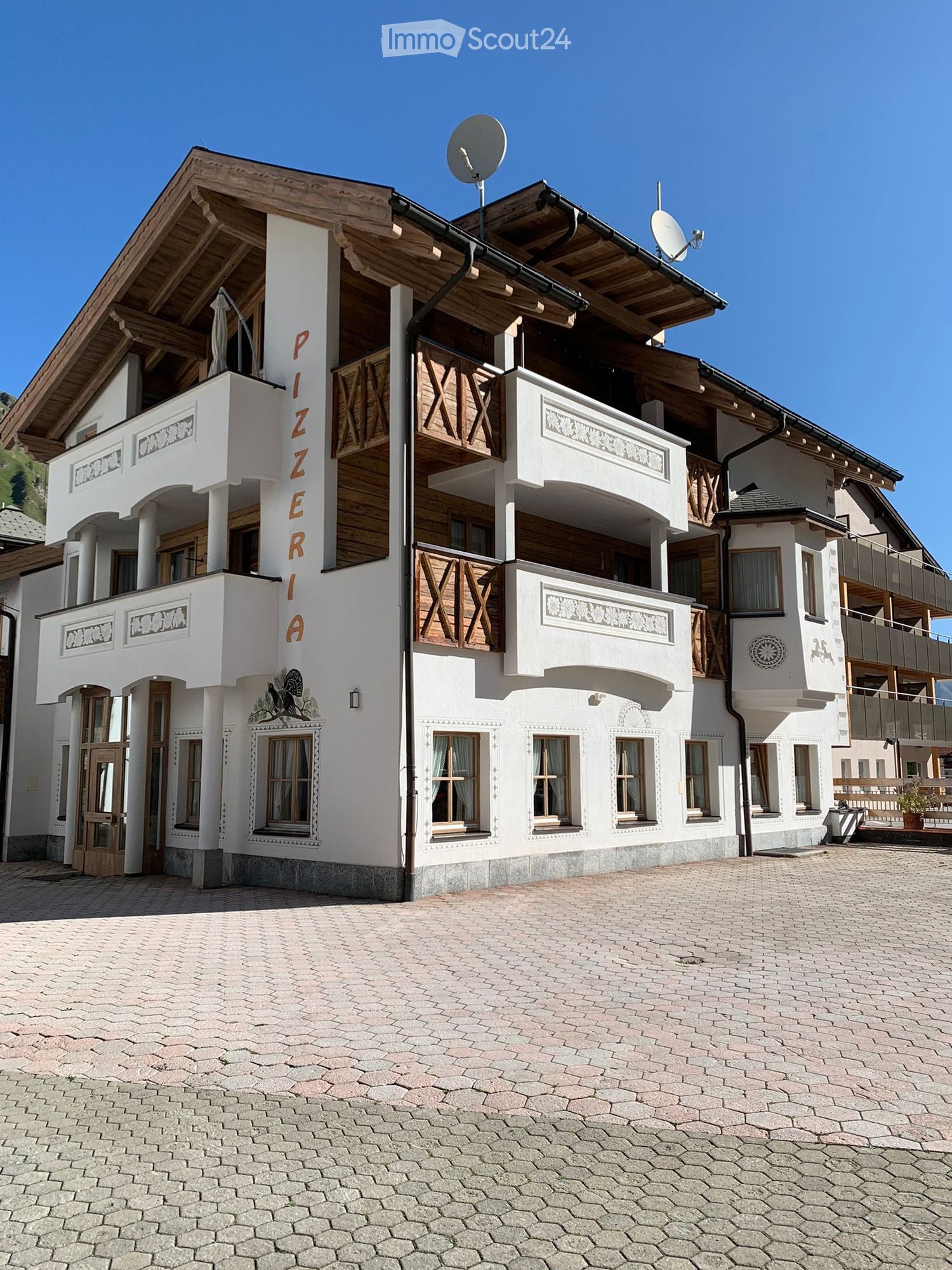 A multi-story building with white walls, wooden accents, and balconies. The building features a sign reading "PIZZERIA" and has multiple windows with curtains. The ground is paved with cobblestones.