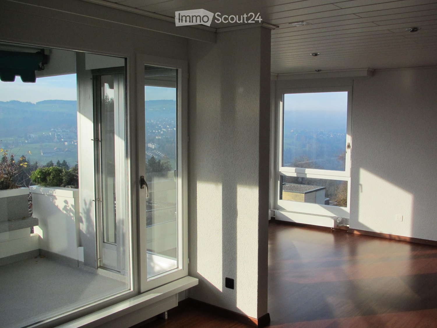 Bright, empty room with large windows and sliding glass doors leading to a balcony, offering scenic views of the landscape.