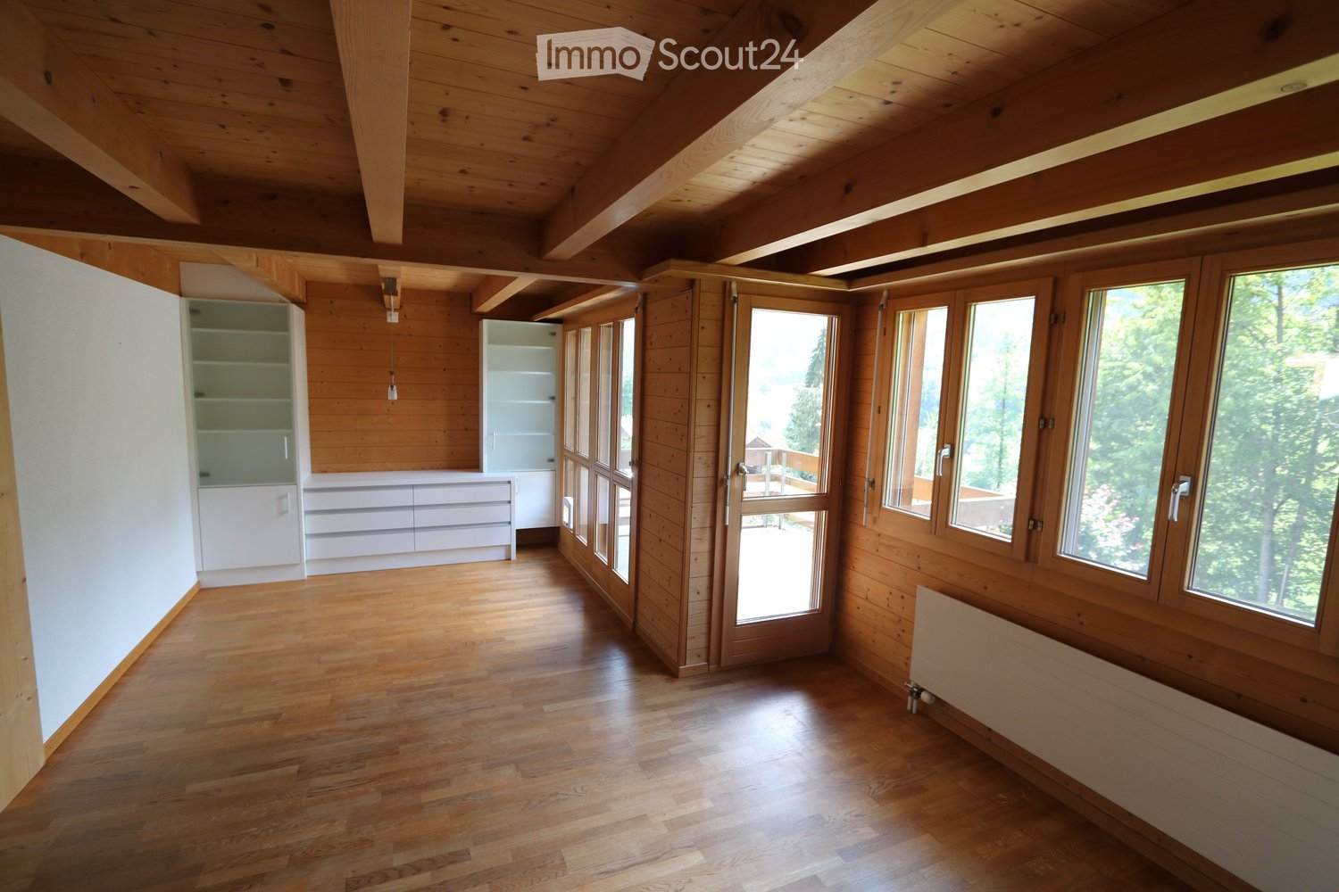 Bright, empty room with wooden walls, ceiling beams, and large windows offering a view of greenery. The floor is made of polished wood, and there are built-in white cabinets and a radiator visible.