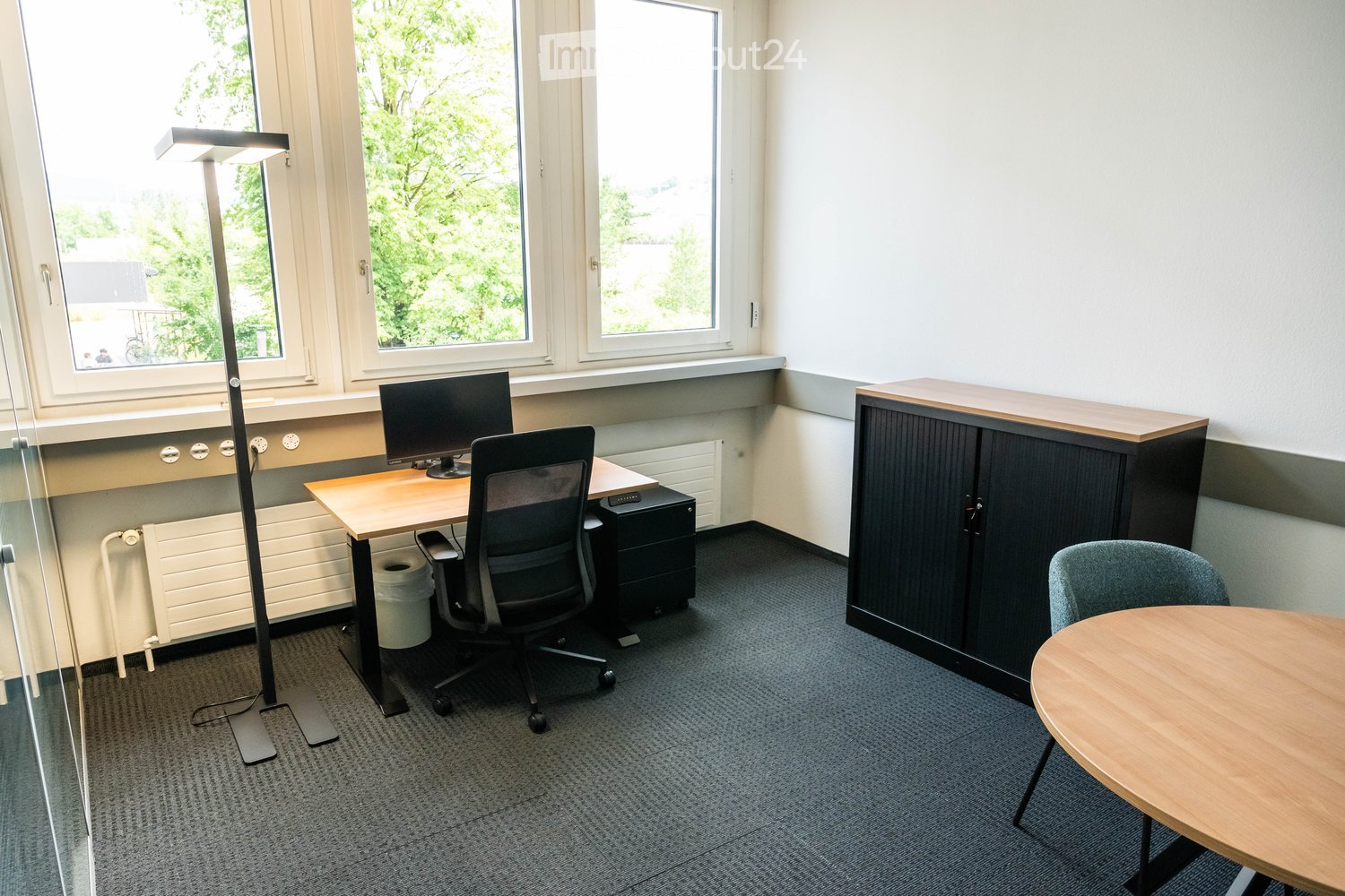 Office space with a desk, chair, computer monitor, cabinet, round table, and floor lamp. Large windows provide natural light.