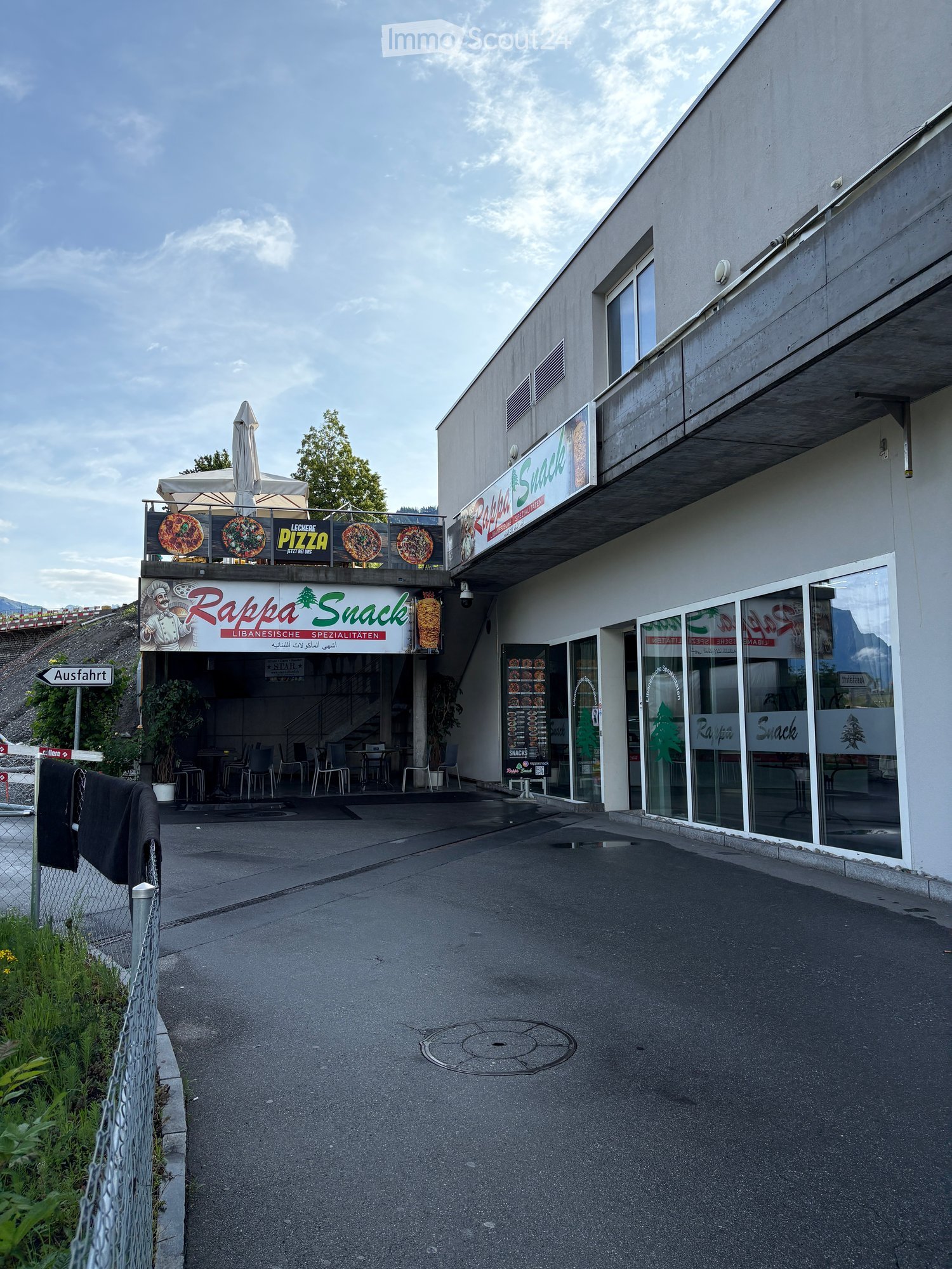 A modern building with a sign reading "Rappa Snack" and a menu board displaying pizza options. The entrance features glass doors and a small outdoor seating area with tables and chairs.