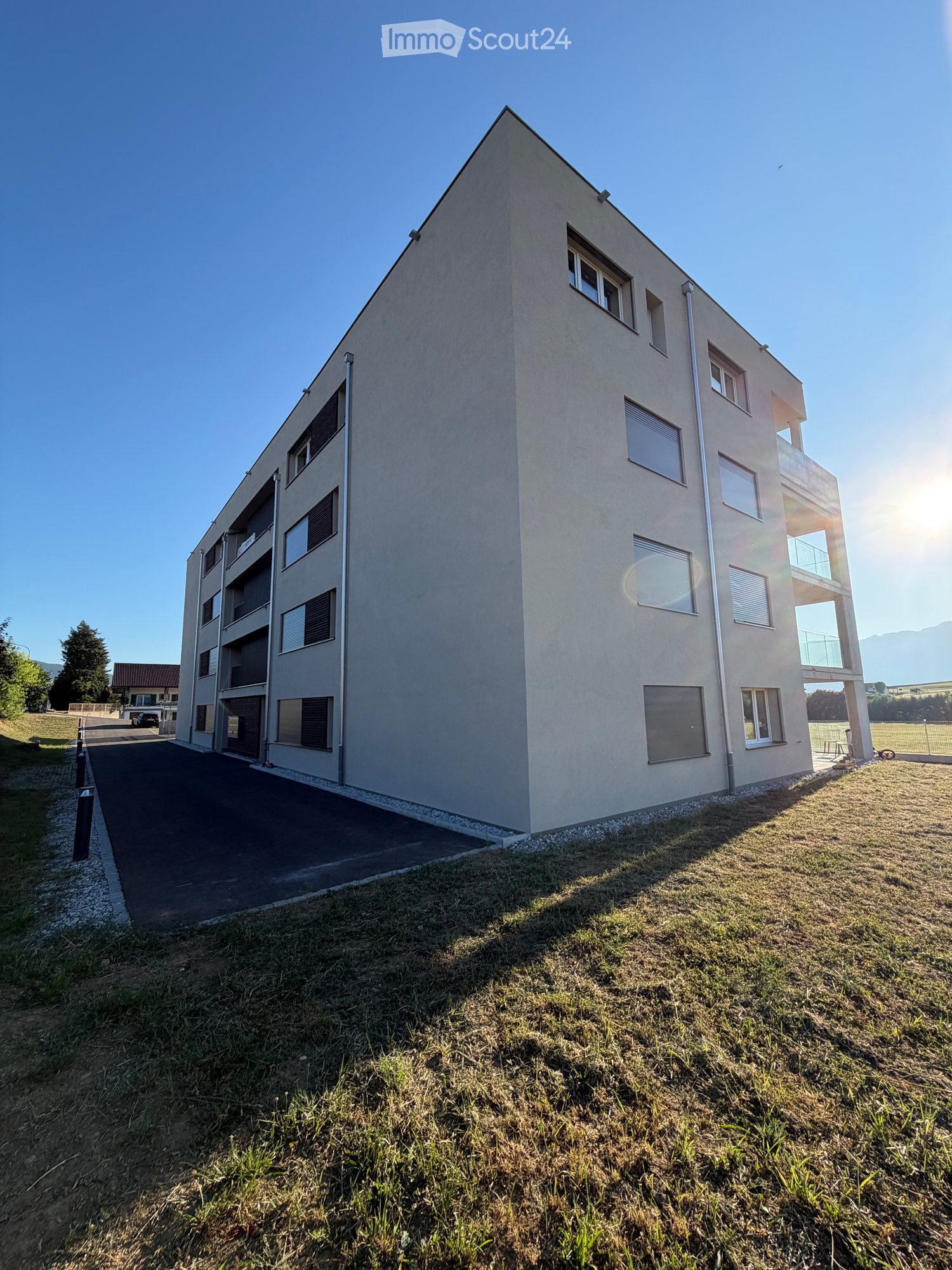 Modern multi-story residential building with beige facade, large windows, and balconies. The building is surrounded by a paved driveway and grassy area.