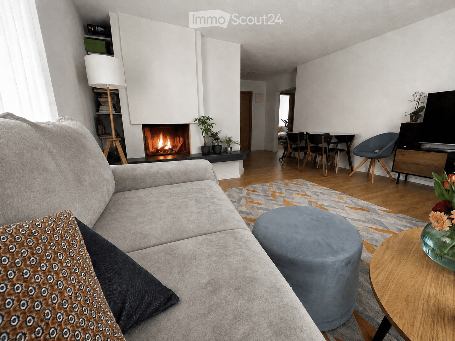 Modern living room with a grey sectional sofa, patterned throw pillow, ottoman, coffee table, floor lamp, fireplace, and dining area with chairs and table.