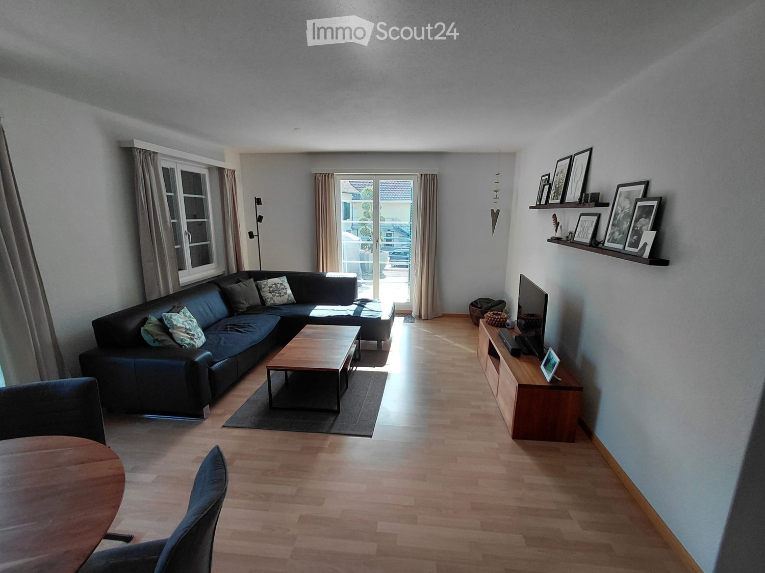 Modern living room with wooden flooring, large window, and sliding glass door leading to a balcony. Features a black sectional sofa, wooden coffee table, TV stand, and shelves with picture frames.