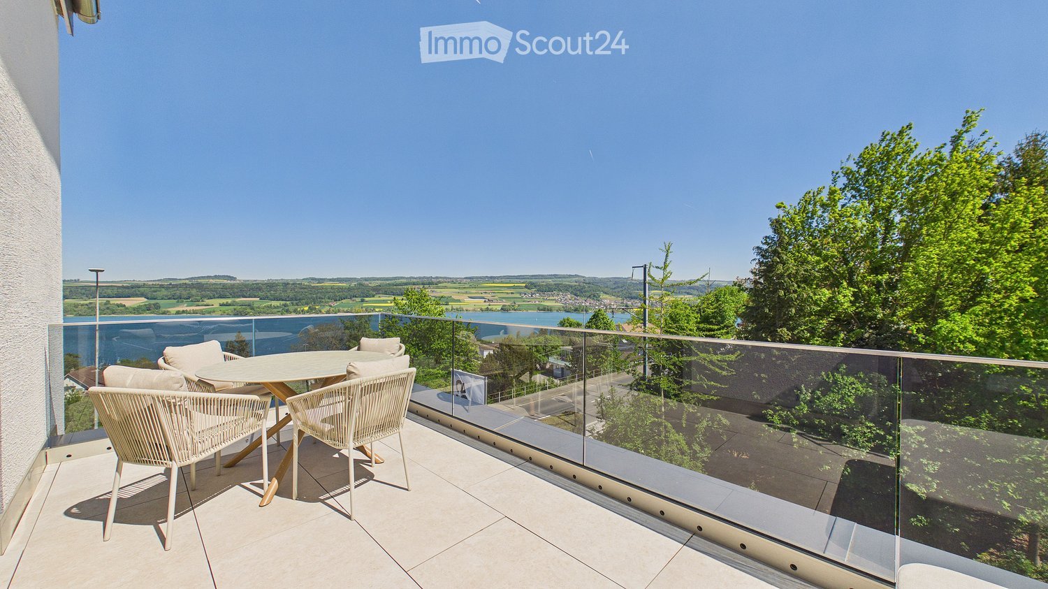 Balcony with a glass railing, overlooking a scenic landscape with a river and hills. Includes a round table and four wicker chairs.