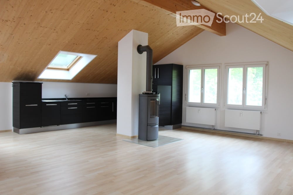 Bright, open room with wooden flooring and exposed wooden beams on the ceiling. Features a modern black kitchen area with cabinets and a sink, a central wood-burning stove with a chimney, and large windows with white frames. The room has a skylight and is