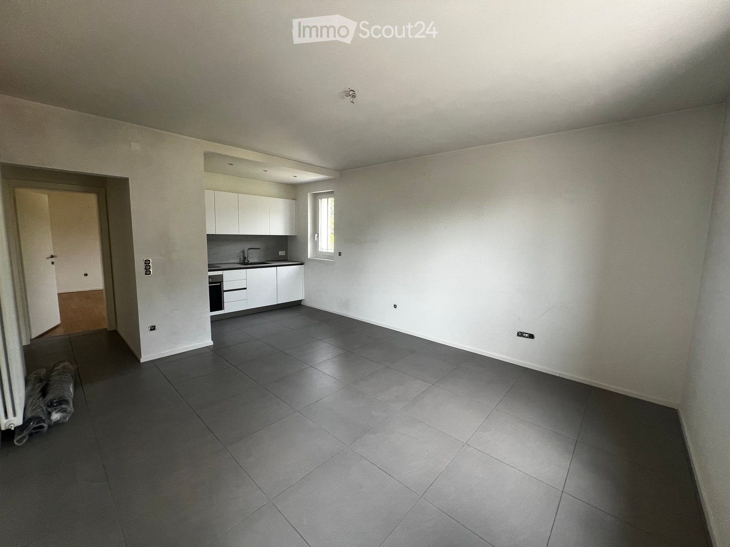 Empty room with grey tiled floor, white walls, and a small kitchen area visible in the background. No furniture or equipment present.