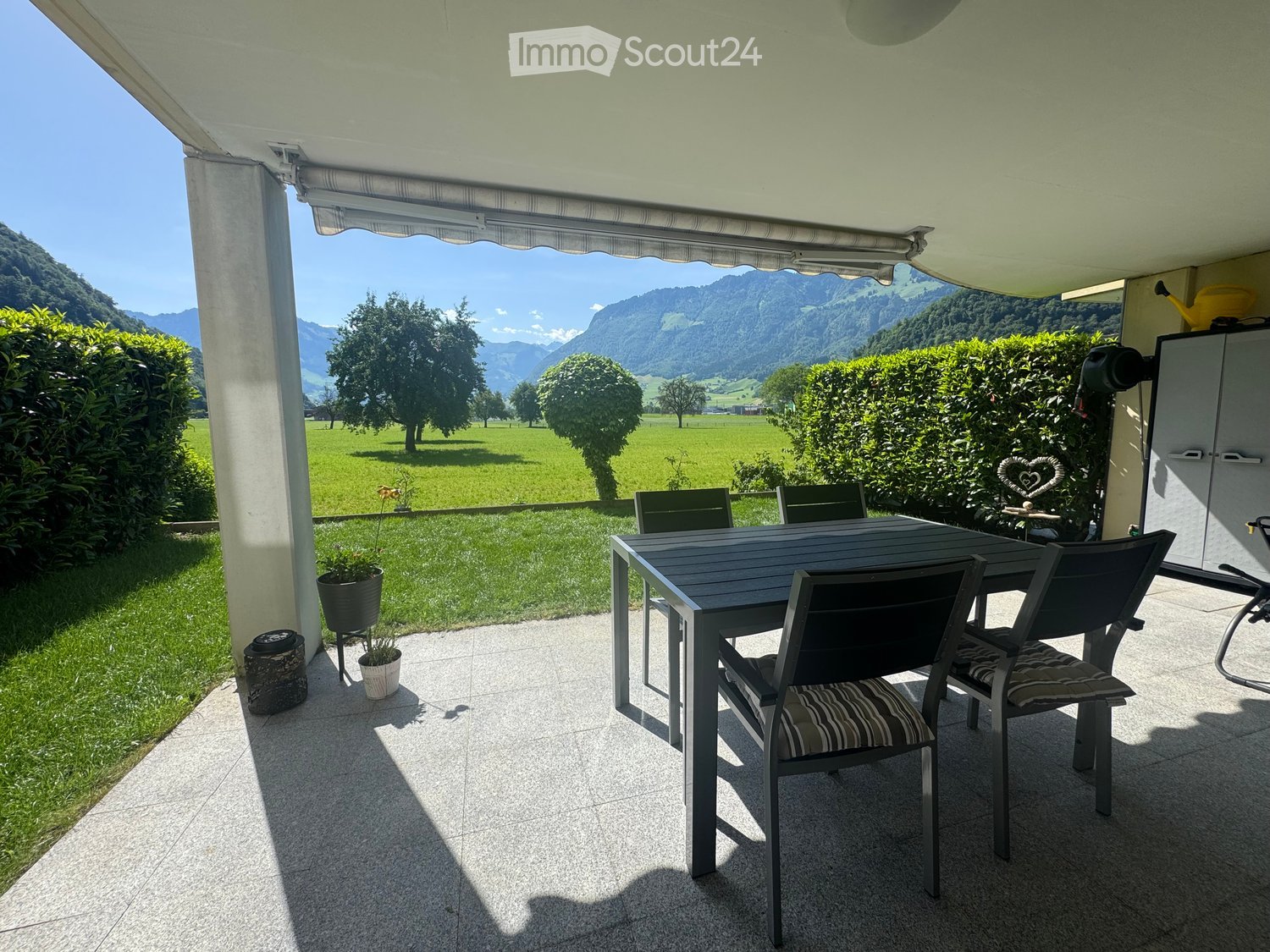 Balcony with a table and chairs, overlooking a green field and mountains.