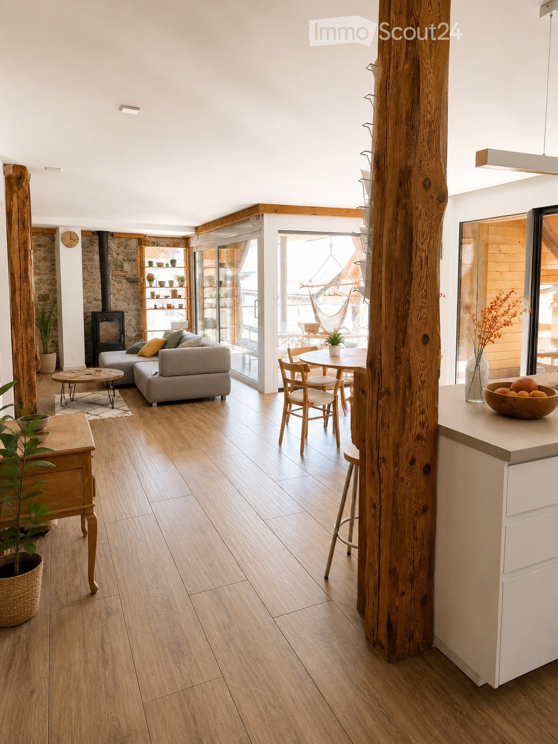 Open-plan living and dining area with wooden flooring, a grey sectional sofa, a small coffee table, a dining table with chairs, a kitchen counter, and a fireplace with a shelf above it.