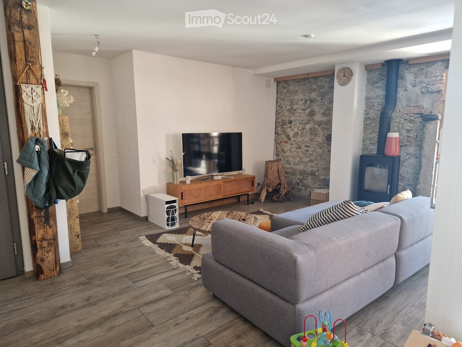 Living room with a grey sofa, coffee table, television, wooden cabinet, wooden stool, fireplace, clock, laptop, and a rug.