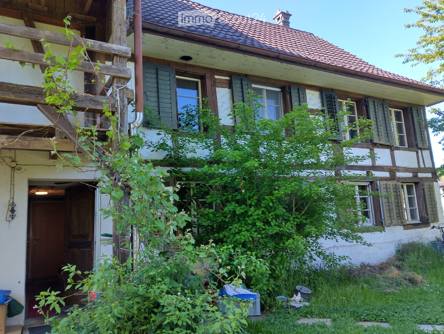 Two-story traditional house with wooden beams, green shutters, and a tiled roof, partially overgrown with vegetation.