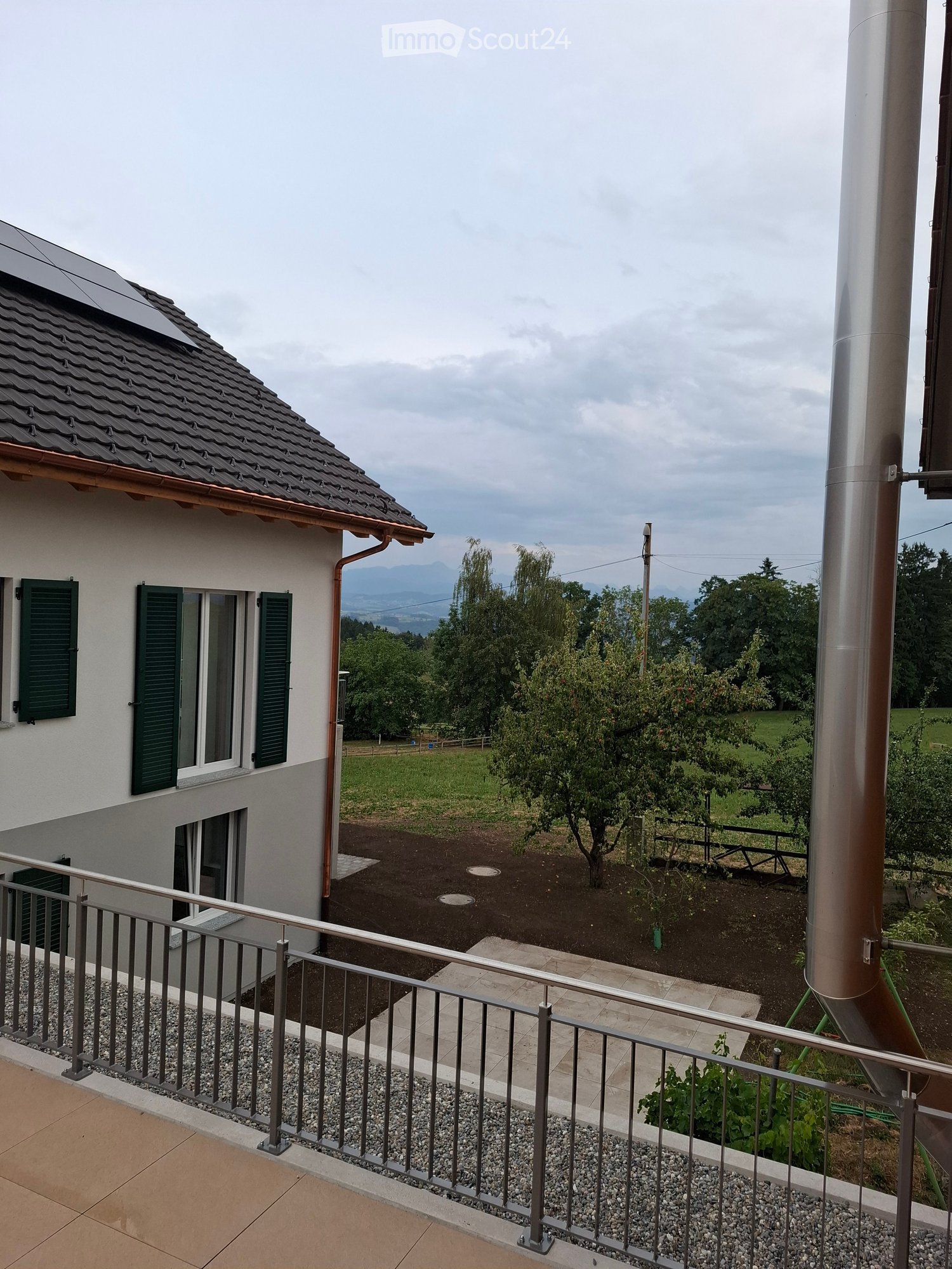 Balcony overlooking a landscaped garden with trees, a house with green shutters, and distant mountains.