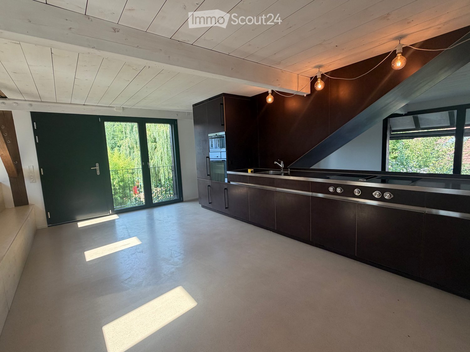 Modern kitchen with dark wooden cabinets, stainless steel appliances, and a large island. The space features a concrete floor and exposed wooden ceiling beams.