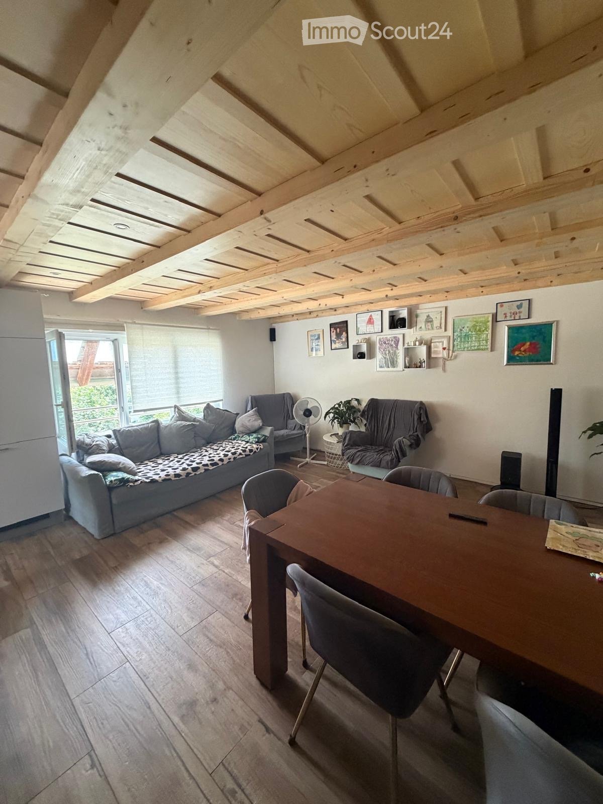 Living room with exposed wooden beams, grey sofa, dining table with chairs, fan, and artwork on the walls.