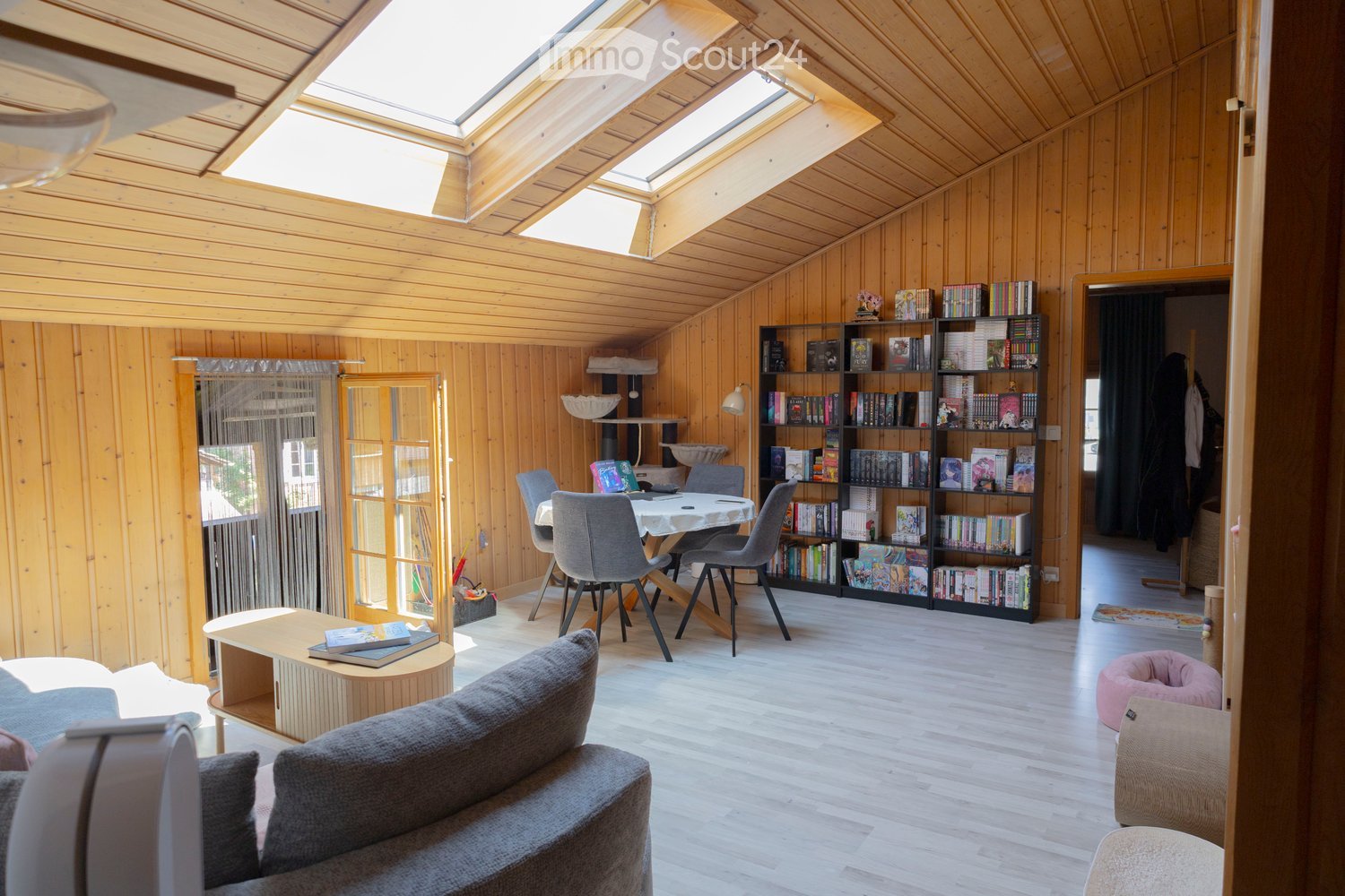 Bright, wooden-paneled room with skylights, featuring a grey sofa, coffee table, bookshelf, dining area with chairs, and a cat tree with a bowl and bed.