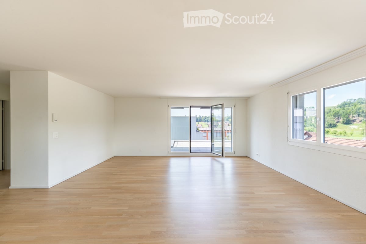 Empty room with light wooden flooring, white walls, large windows, and a set of sliding glass doors leading to a balcony.