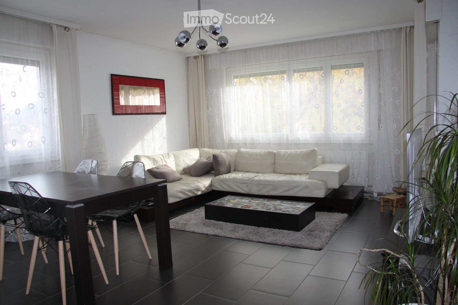 Modern living room with white sectional sofa, coffee table, dining table with chairs, floor lamp, and large windows with sheer curtains.