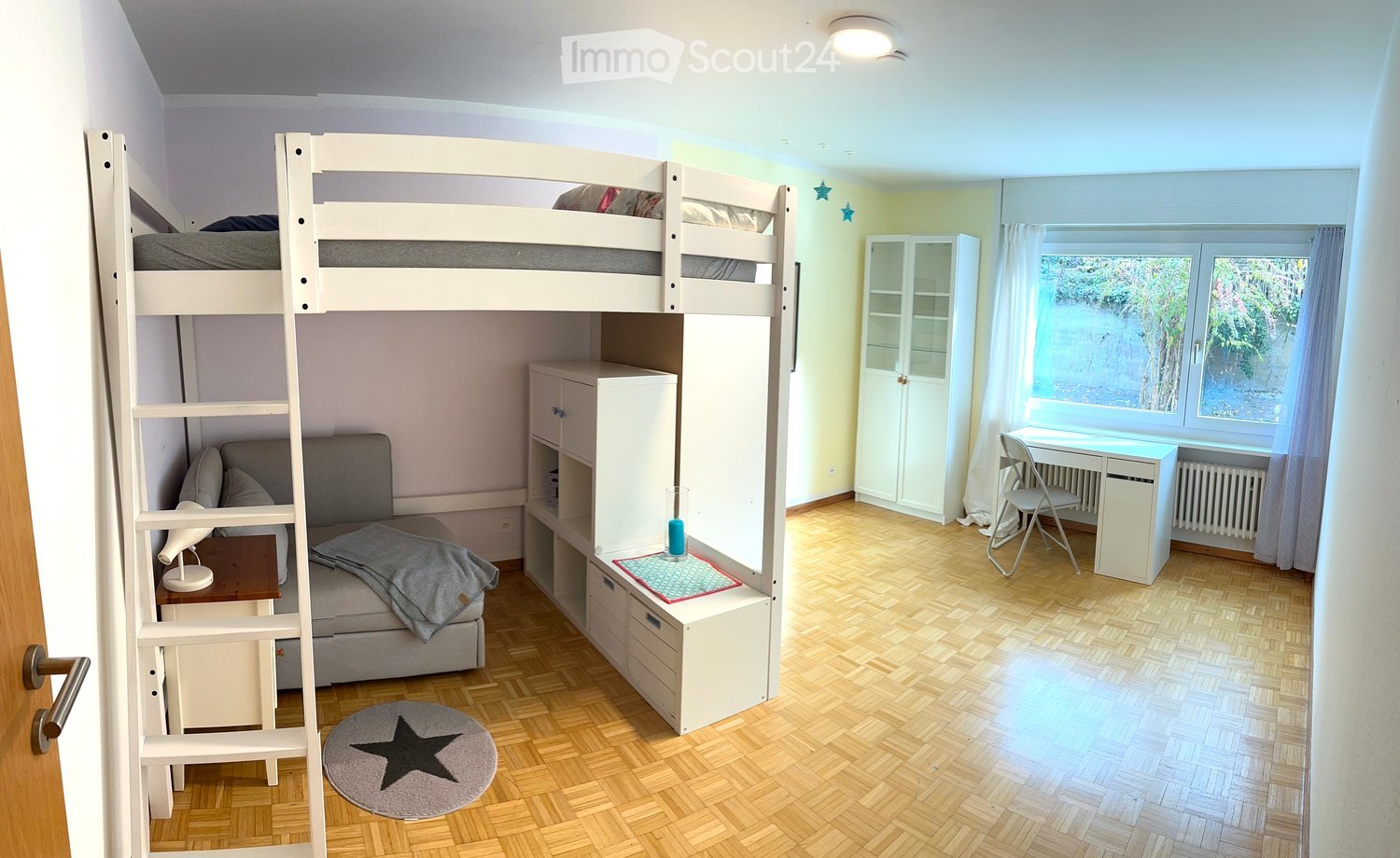 A well-lit bedroom with a bunk bed, a small sofa, a desk, and a chair. The room features wooden flooring, white walls, and large windows with curtains.