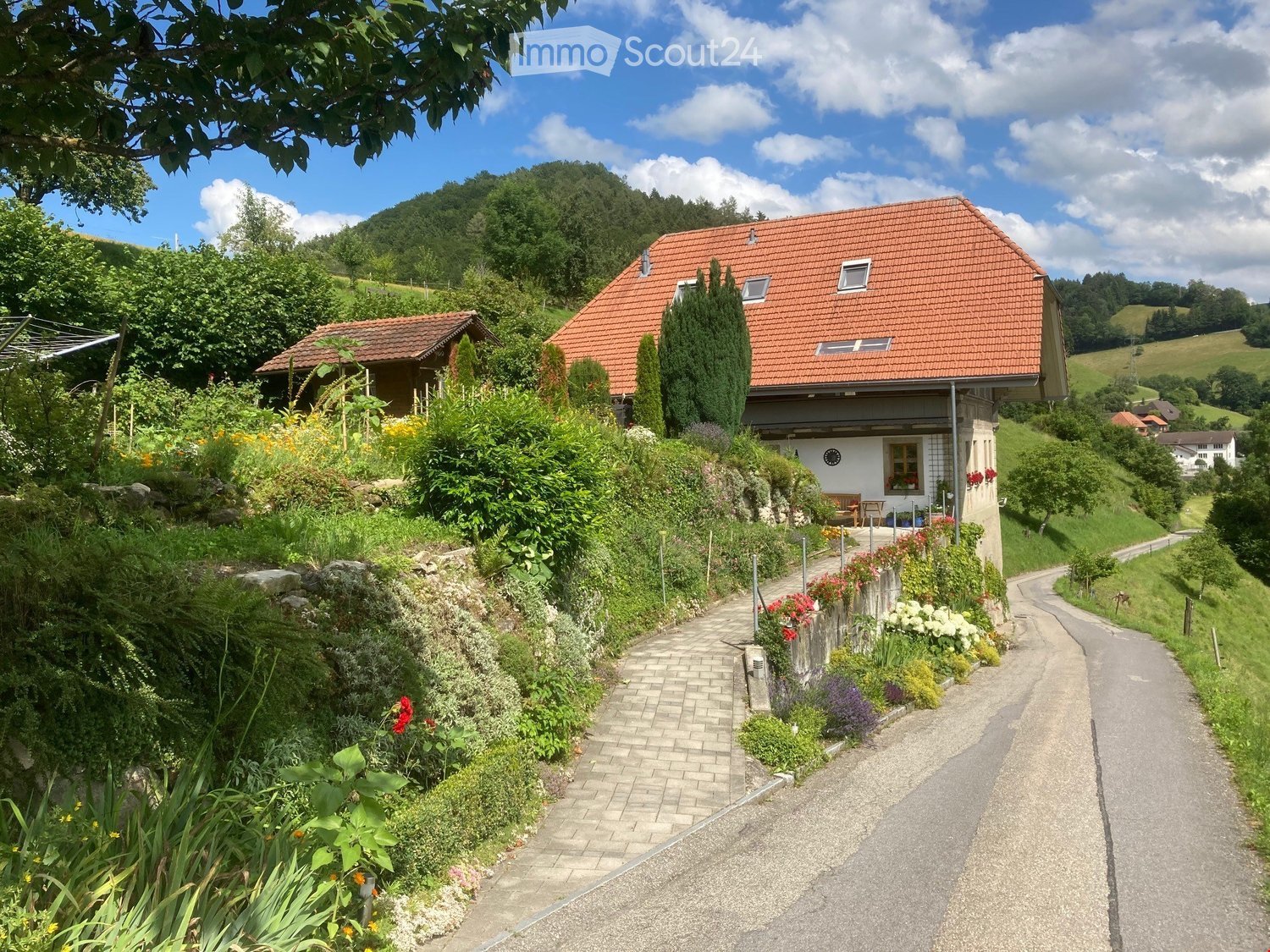 Two-story house with red-tiled roof, surrounded by lush garden and greenery, situated in a hilly landscape with other houses visible in the distance.
