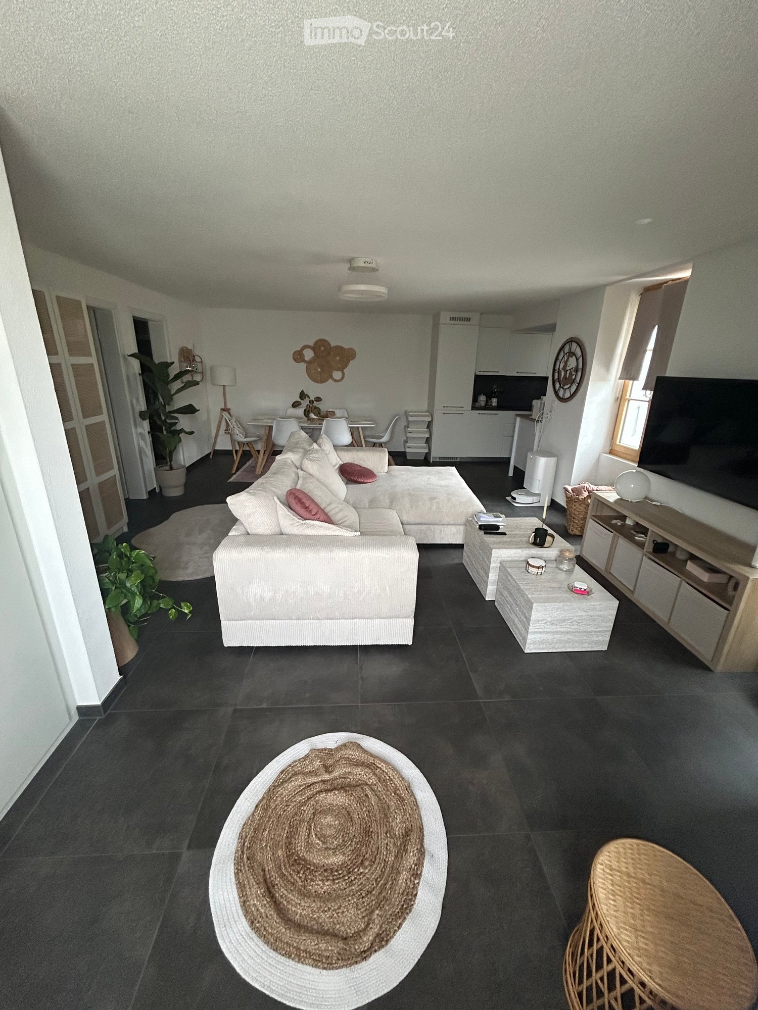 Modern living room with a white sectional sofa, coffee table, side tables, dining area with chairs and table, TV, floor lamp, and plants.