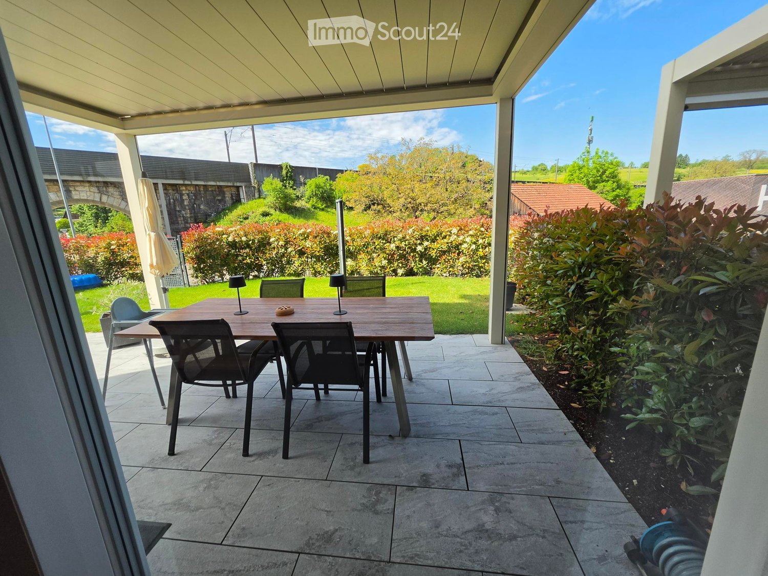 Patio area with a wooden table, chairs, and outdoor lighting, surrounded by greenery and a clear sky.