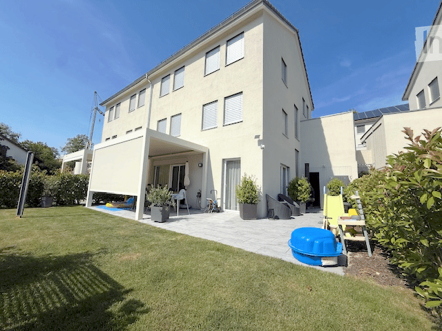 A modern two-story house with a spacious garden and patio area. The patio features a covered seating area with outdoor furniture, potted plants, and a small swimming pool. The lawn is well-maintained, and the house has large windows and a clean, contempor
