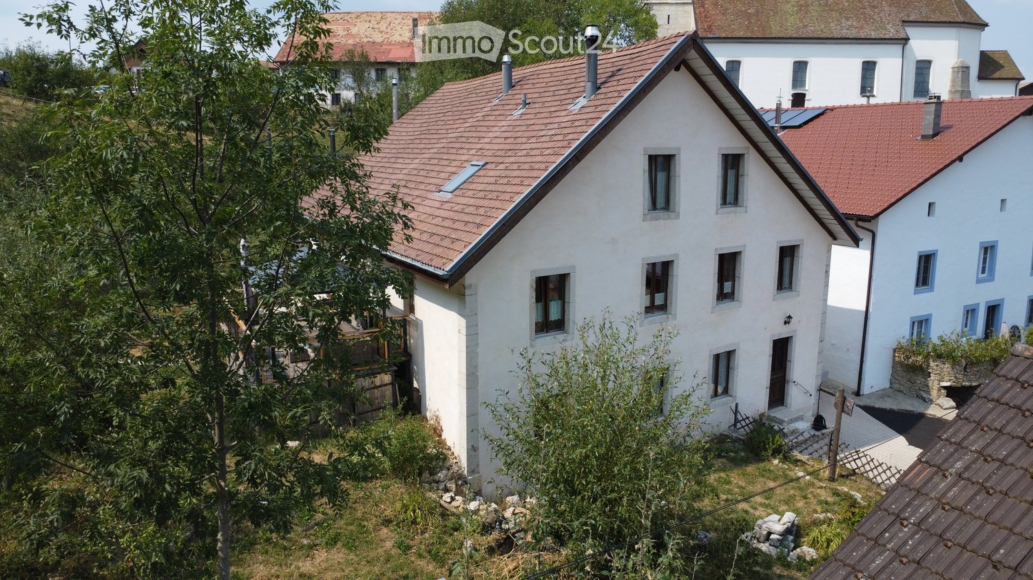 A two-story white house with a red-tiled roof, multiple windows, and a small garden area in front.
