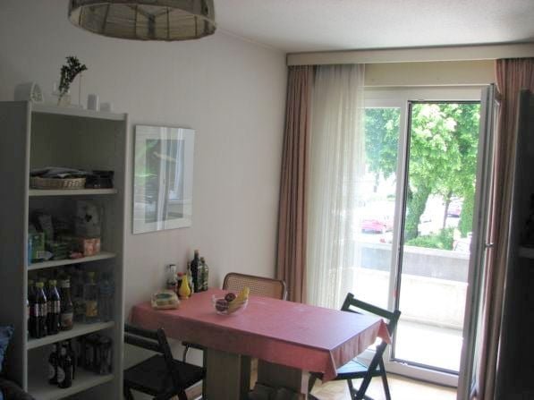 Dining area with a table covered in a pink tablecloth, surrounded by chairs. A fruit bowl with bananas and other fruits sits on the table. A tall cupboard with various bottles and items is visible on the left side. A large window with curtains lets in nat