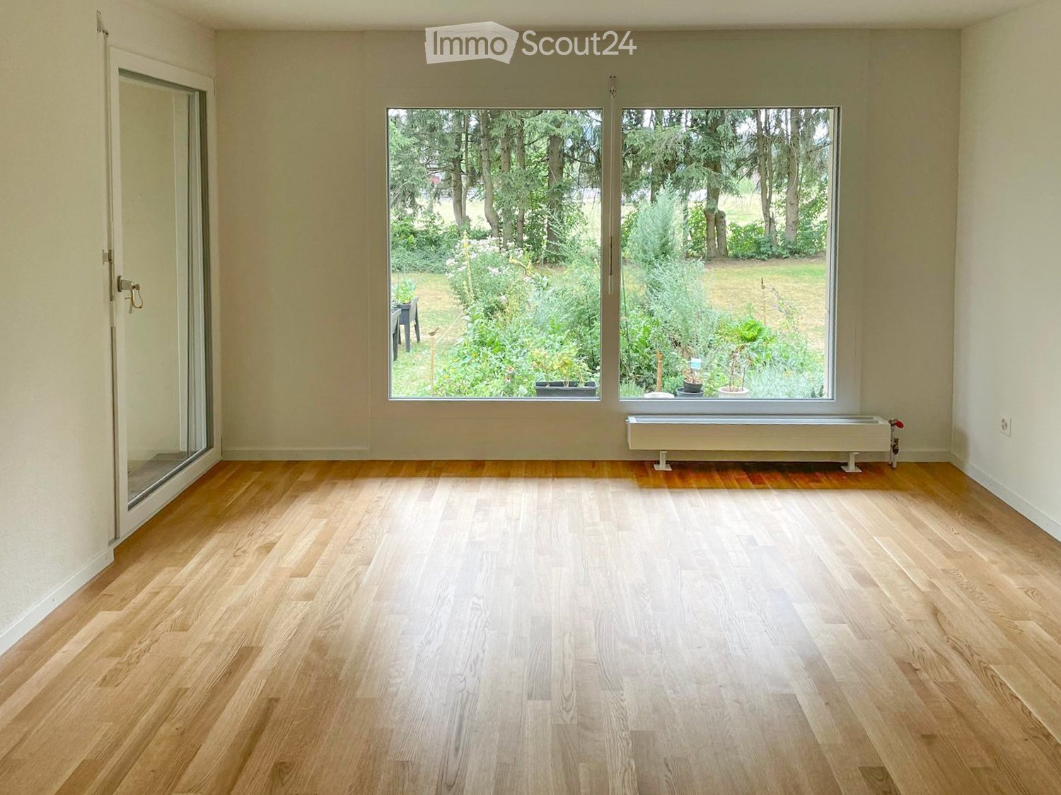 Empty room with light wooden flooring, large windows overlooking a garden, and a sliding glass door leading outside.