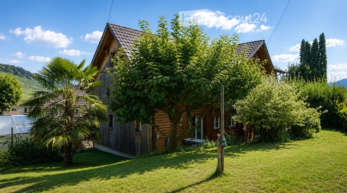 A wooden house with a tiled roof, surrounded by lush greenery and trees, including a palm tree in the front yard.