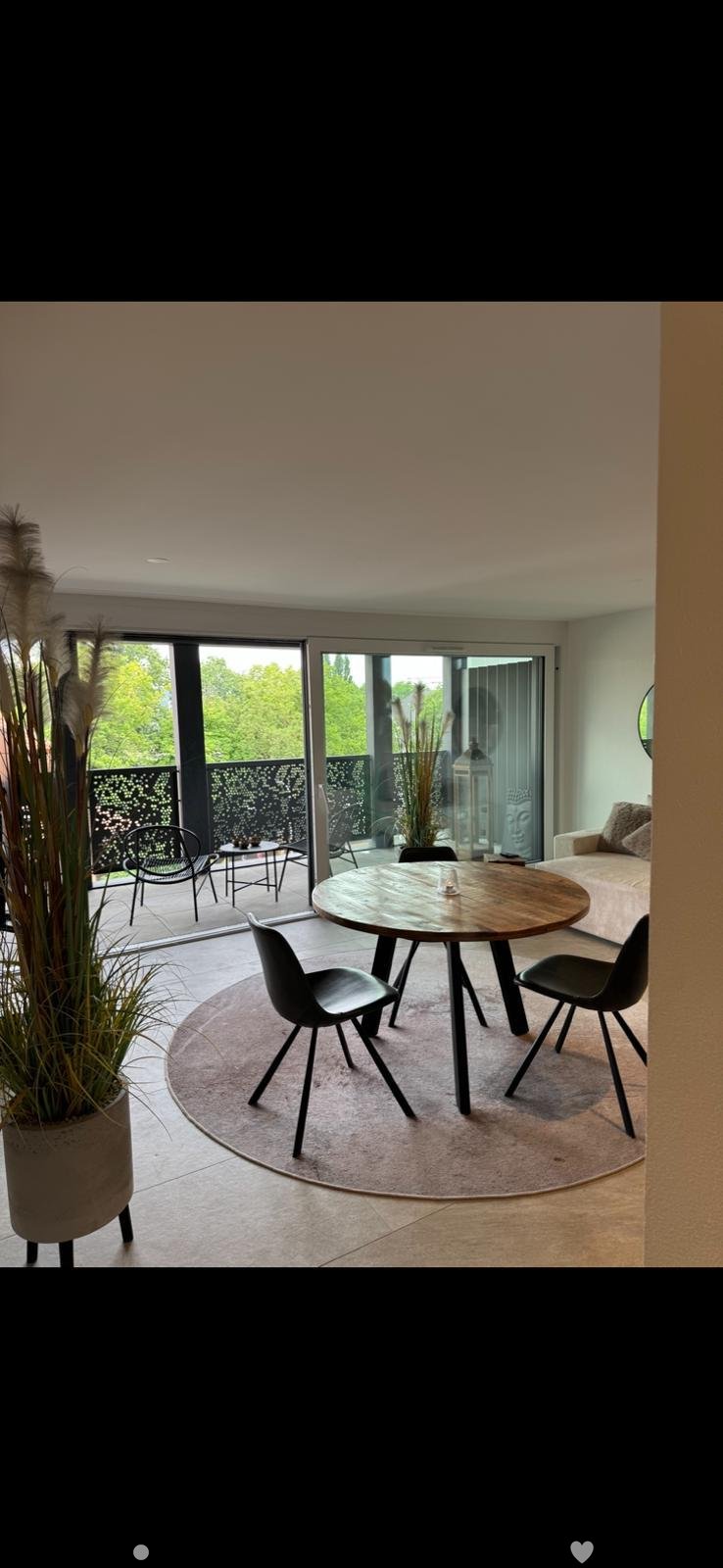 Modern dining area with a round wooden table, black chairs, a sofa, and a small side table, connected to a balcony with outdoor seating.