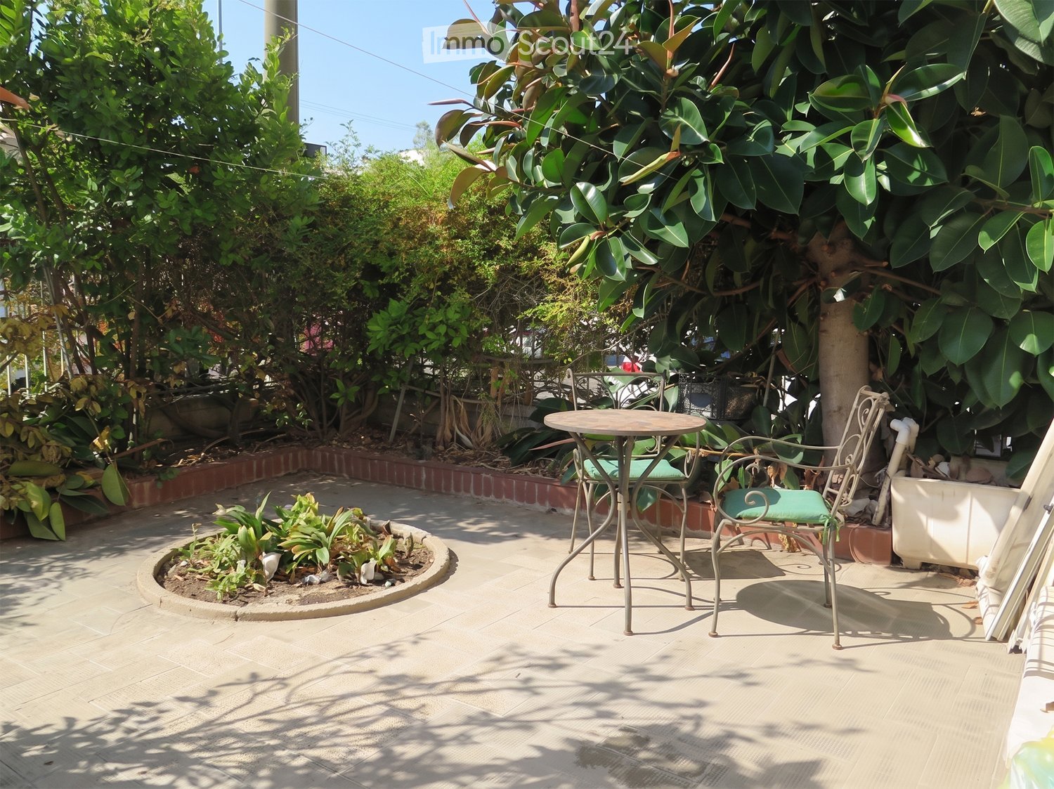 A small outdoor patio with a round table and two chairs, surrounded by lush greenery and plants.