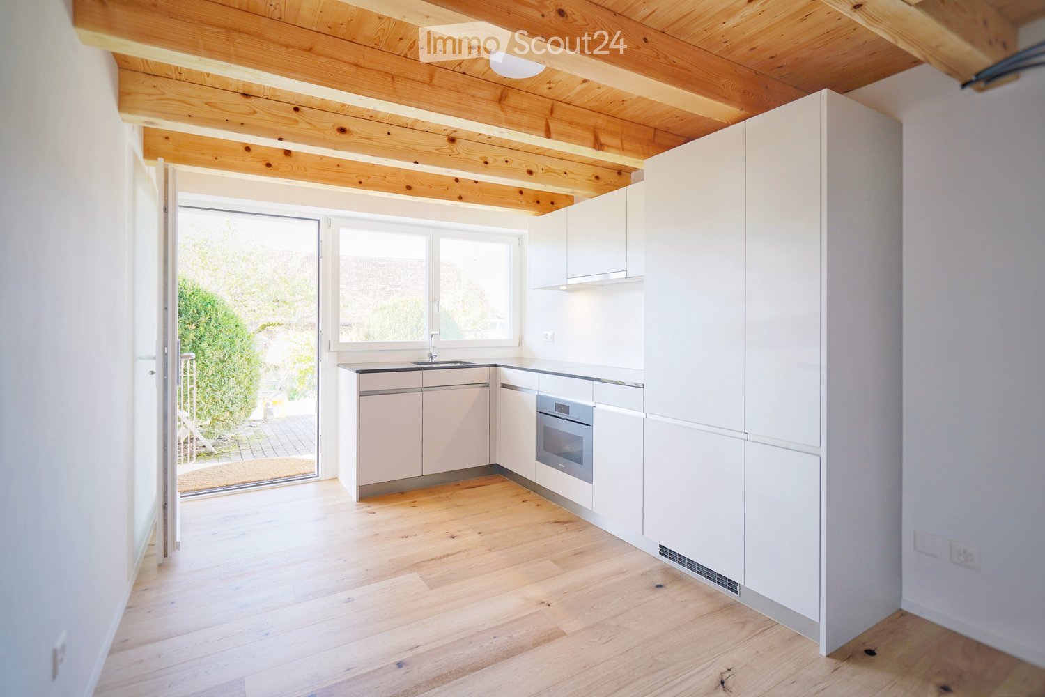 Modern kitchen with white cabinets, wooden flooring, and a visible oven. Large windows and a sliding door lead to an outdoor area.