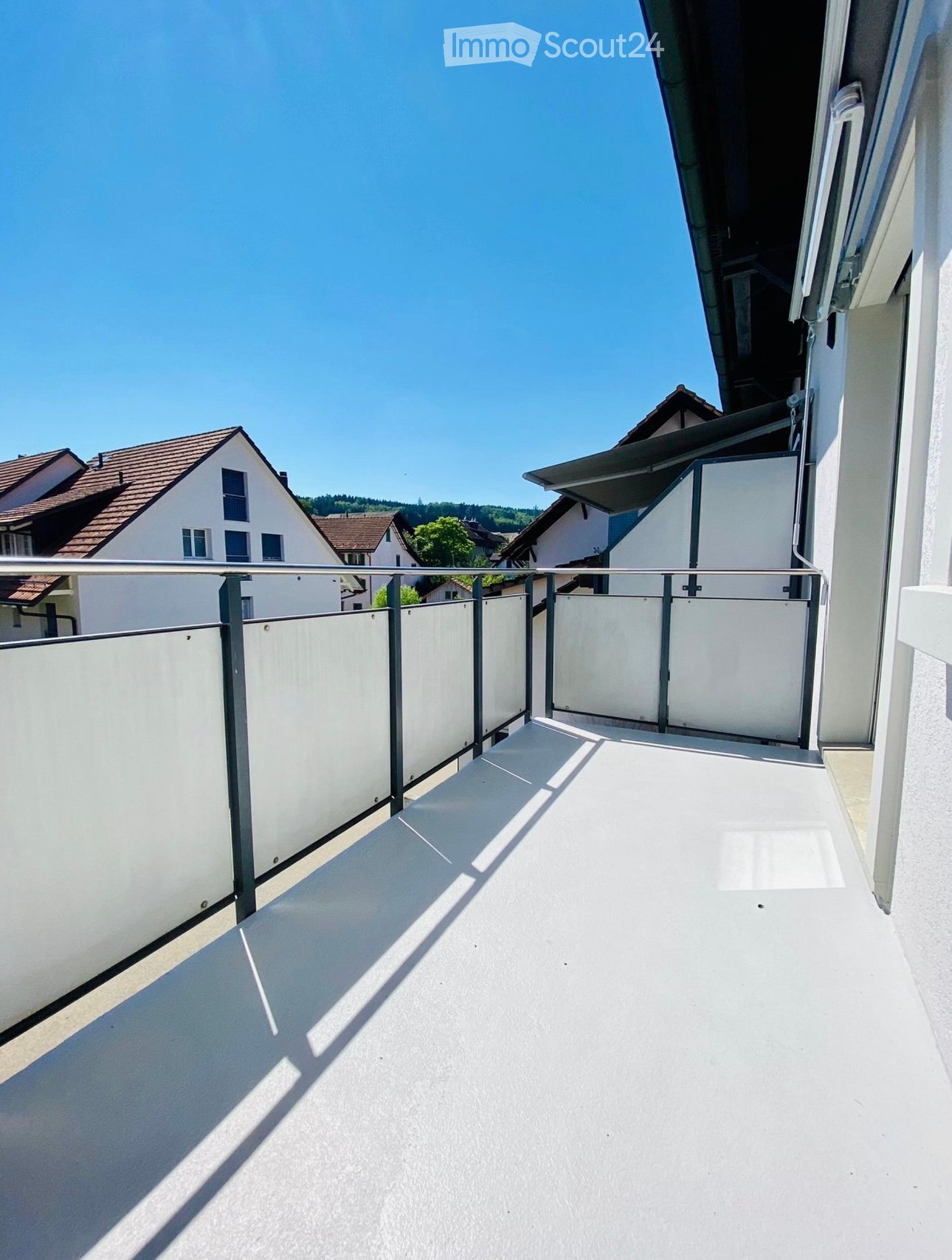 Balcony with white floor, metal railing, and white privacy screens, overlooking a residential area with houses and trees.