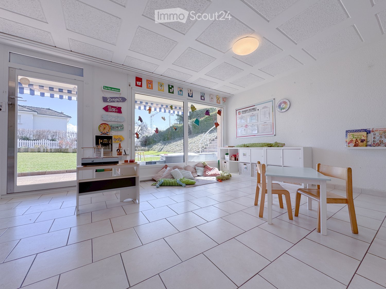 Bright, airy room with white tiled flooring, large windows, and a skylight ceiling. Contains a small table with chairs, a cabinet, a screen, and various toys and decorations.